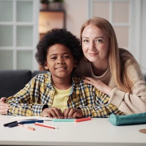 Portrait of young foster mother looking at camera with her adoptive son while they sitting at table at home